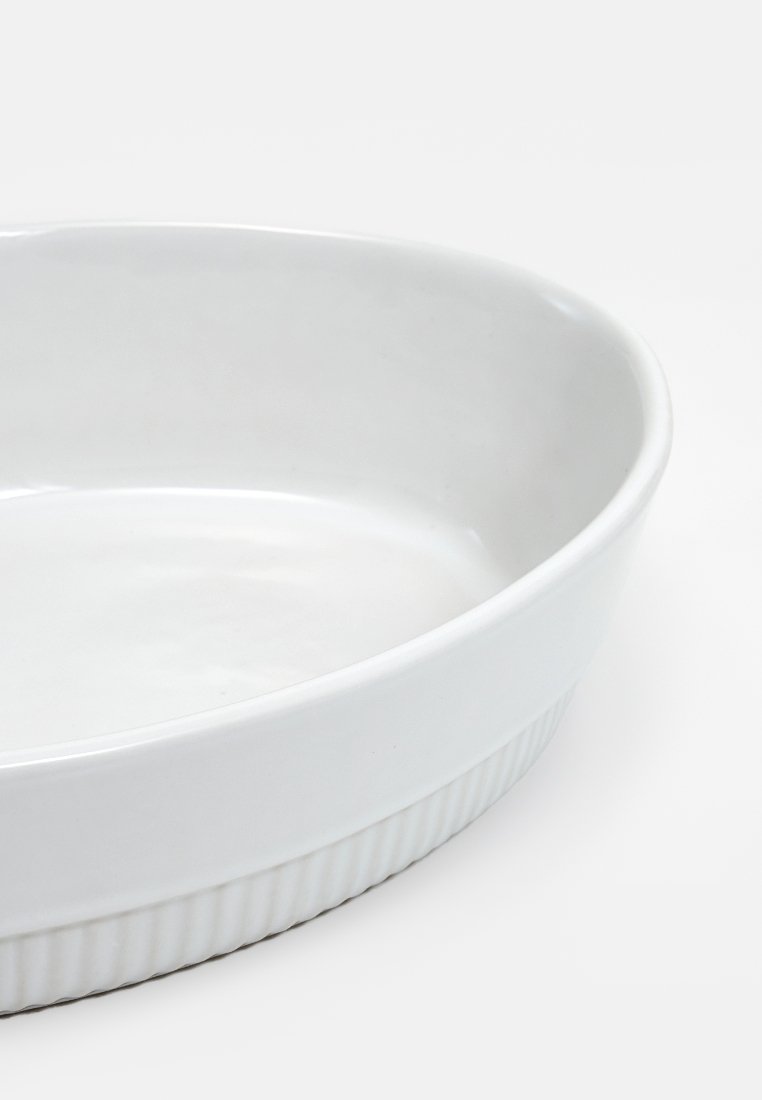 Pirofila in ceramica bianca con superficie liscia, forma ovale e accentature rigate lungo i lati. Adatta per uso in forno.