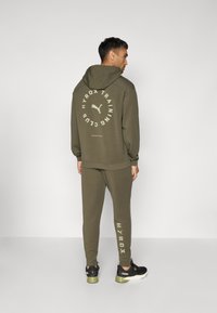Olivgrünes Sweatpants- und Hoodie-Set mit weißem Logo und Text auf dem Rücken und am linken Hosenbein. Schwarze Sneakers mit grünen Akzenten.