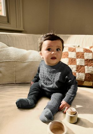 Grå tekstureret strikket outfit med ribbede detaljer. Barnet sidder på en beige sofa, med trælegetøj i nærheden og en ternet pude i brun og creme.