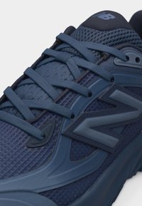 Κοντινό πλάνο ενός σκούρου μπλε αθλητικού παπουτσιού New Balance που δείχνει το πλεκτό επάνω μέρος από δίχτυ, τις λεπτομερείς κορδόνια και το μεγάλο λογότυπο "N" στη πλευρά.