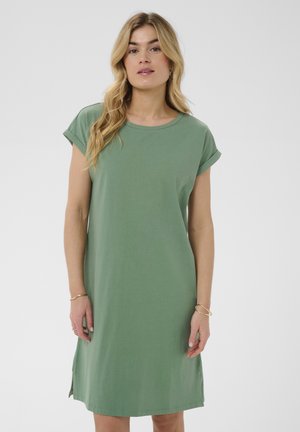 Frau mit langen blonden Haaren, die ein einfaches grünes knielanges Kleid mit kurzen Ärmeln und goldene Armbänder an beiden Handgelenken vor einem schlichten Hintergrund trägt.