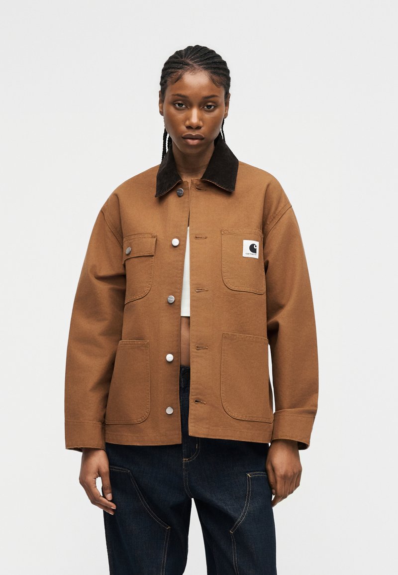 Donna che indossa una giacca marrone con bottoni, colletto nero, due tasche sul petto e due tasche inferiori, sopra una maglia bianca e pantaloni scuri.