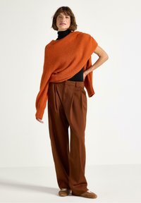 Kvinna iklädd en bränd orange oversized tröja över svart polo, bruna vida byxor och beige sneakers, stående mot en vit vägg.