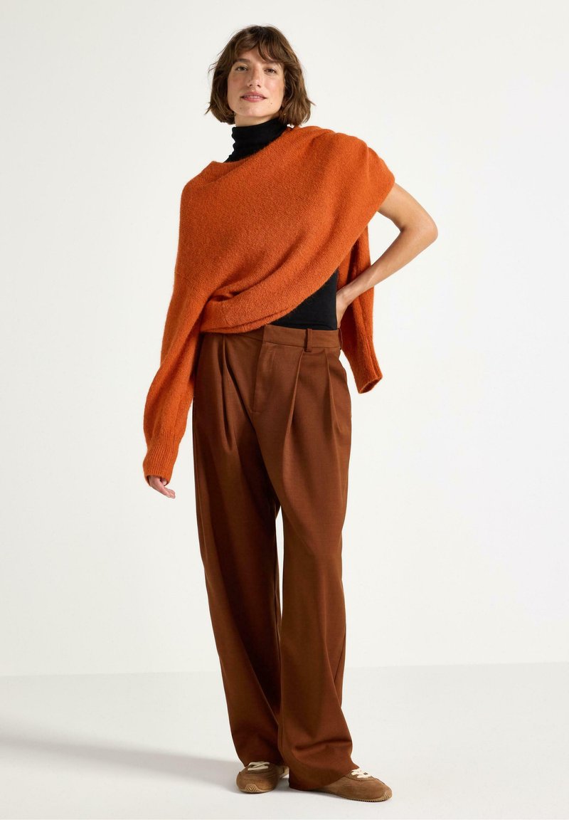 Kvinna iklädd en bränd orange oversized tröja över svart polo, bruna vida byxor och beige sneakers, stående mot en vit vägg.