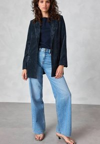 Kvinna klädd i en mörk marinblå kordjacka, marinblå topp, ljusblå vida jeans och öppna klacksandaler, stående mot en grå bakgrund.
