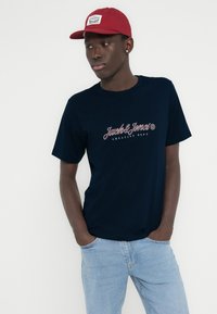 Μαύρο T-shirt με λογότυπο "Jack & Jones" σε λευκό και κόκκινο γραφικό, συνδυασμένο με τζιν παντελόνι ανοιχτού μπλε και κόκκινο καπέλο με λογότυπο.