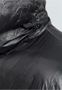 Sort pufferjakke med en skrumpet tekstur, der har en lynlåslukning ved halsen og en glat inderside, hvilket fremhæver det lette design.