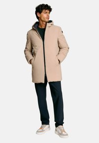 Uomo in piedi con le mani nelle tasche, indossa un cappotto invernale beige con cappuccio, pantaloni neri e sneakers grigie con dettagli arancioni.