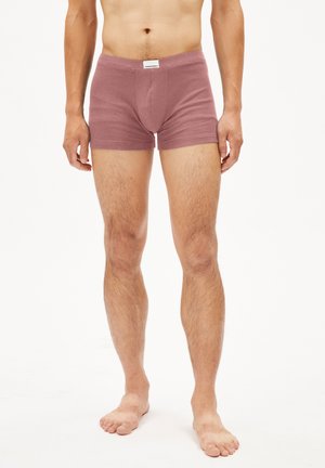 Homme debout pieds nus portant un caleçon mauve avec un ceinturon étiqueté sur fond blanc.