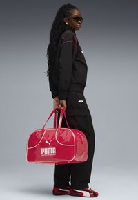 Pink glossy duffel bag with white accents and the Puma logo, paired with a black outfit and red-accented footwear.