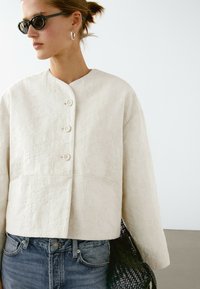 Kvinde iført en hvid tekstureret knapjakke, blå jeans, sorte ovale solbriller, sølv hoop øreringe og holder en sort vævet taske.