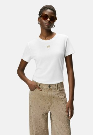 Donna che indossa grandi occhiali da sole tartarugati, camicia bianca a maniche corte con piccolo logo e pantaloni beige larghi con borchie, posa con una mano in tasca.