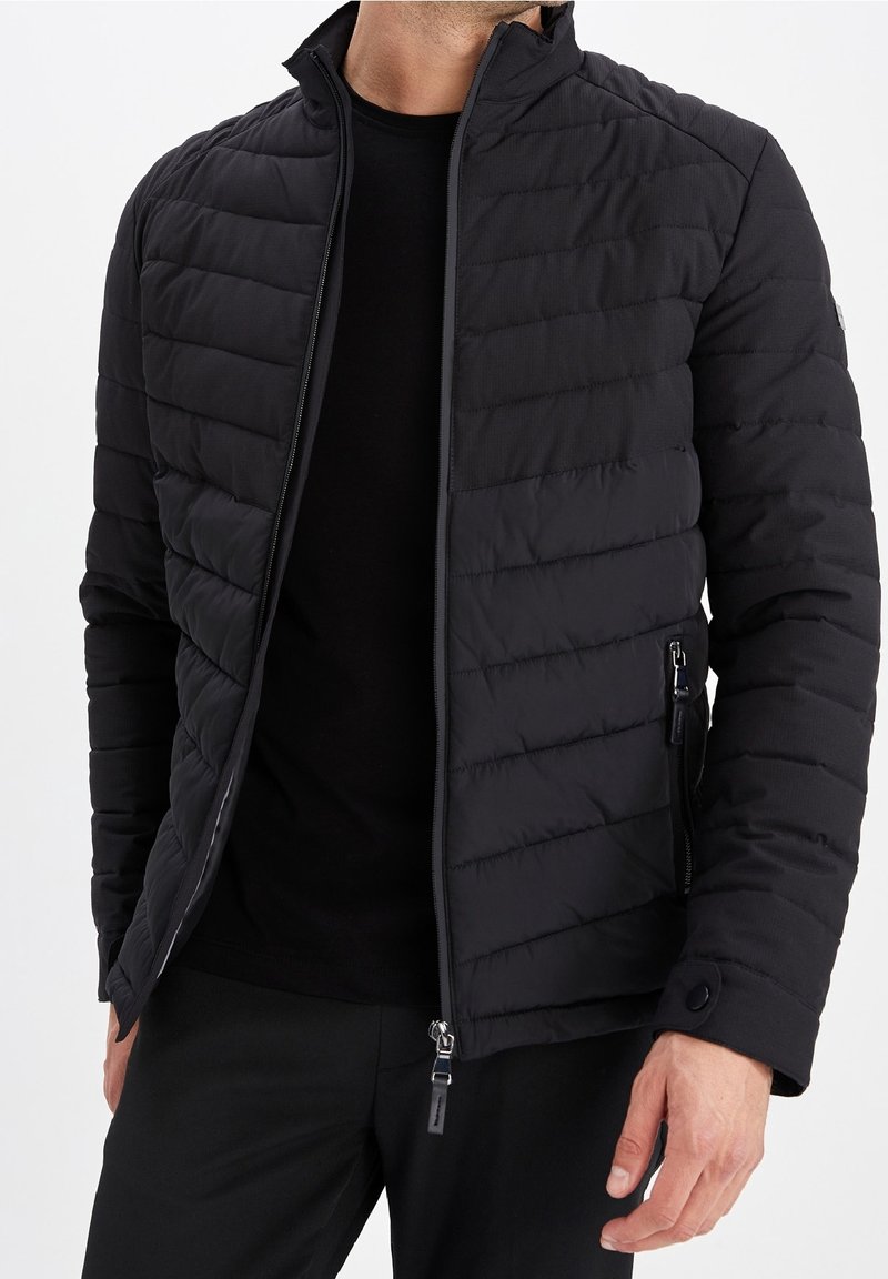 Homme portant une veste matelassée noire zippée et un pantalon noir, debout devant un fond clair uni.