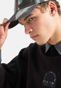 Cappello mimetico con visiera strutturata, motivo grigio e teal, e un'etichetta con logo; abbinato a una felpa nera con logo.