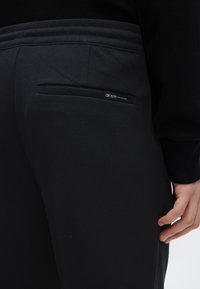 Μαύρο τζιν παντελόνι με πίσω τσέπη που φέρει ετικέτα με εμπορικό σήμα. Το ύφασμα έχει υφή με σχέδι ο, ενώ η ζώνη μέσης είναι λεία.