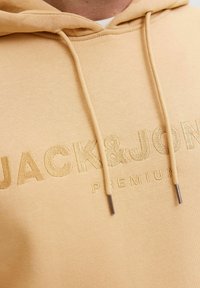 Sudadera beige con cordón y logo en relieve "JACK&JON". Textura suave y ajuste casual, con extremos de metal en los cordones.