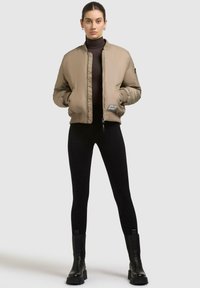 Beige Bomberjacke mit Reißverschluss, gerippten Bündchen und Saum, kombiniert mit schwarzen Leggings und Stiefeletten. Glatte Textur mit minimalen Details.