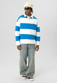 Polo à manches longues avec des rayures horizontales bleues et blanches, en coton ; porté avec un jean bleu clair et des baskets noires ; accessoirisé d'un bonnet beige.