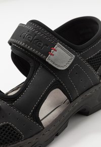 Svart sandal i mesh och slät läder, justerbar rem, grå accenter och röd sömnad. Har utskärningar och en robust sula.