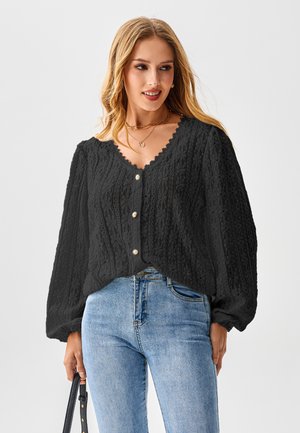 Schwarze Strickjacke mit strukturierten Mustern, gerüschtem Ausschnitt und Perlenknöpfen. Kombiniert mit hellblauen, hoch taillierten Jeans.