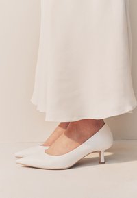 Hvide læderhøje hæle med en spids tå og en slank hæl, kombineret med et flydende hvidt stofskørt.