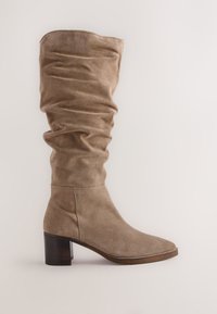 Stivale alto fino al ginocchio in camoscio beige con design morbido, punta quadrata e tacco a blocco in legno. Materiale testurizzato e pieghe morbide sono evidenti.