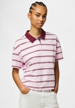 Ung kvinde med flettet hår iført en polo-shirt med lyserøde og mørkerøde striber og hvide bukser, hænderne i lommerne, neutral baggrund.