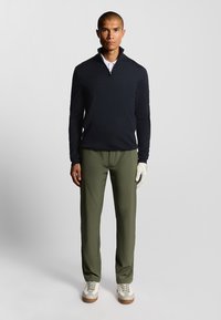 Maglione con zip navy, pantaloni verde oliva, sneakers bianche con suola texturizzata e guanto da golf bianco indossato sulla mano sinistra.