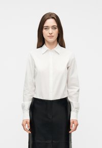 Γυναίκα με ίσια καστανά μαλλιά που φοράει λευκό πουκάμισο με κουμπιά και μαύρη δερμάτινη φούστα, στέκεται μπροστά σε απλό φωτεινό φόντο.