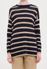 Person, der einen langärmligen Pullover mit horizontalen Streifen in Marineblau und Beige sowie beigen Hosen trägt, steht vor einem schlichten hellen Hintergrund.