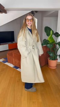 Jovem mulher vestindo um trench coat bege com cinto e óculos de sol, em pé na sala de estar com um sofá laranja, chão de madeira e uma grande planta em vaso.