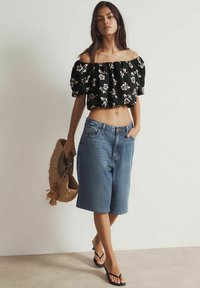 Zwarte off-the-shoulder bloemen crop top gecombineerd met licht denim knielange shorts, een geweven strooien tas dragend en zwarte slippers.