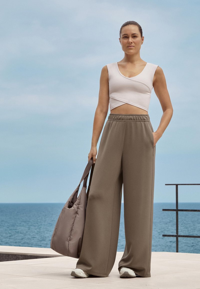 Bijeli crop top s uvijenim dizajnom, uparen s lako prilagođenim hlačama u maslinasto zelenoj boji. Ima mekanu, teksturiranu tkaninu i sivu torbu preko ramena.