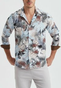 Chemise à manches courtes présentant un motif floral en bleu, beige et marron sur fond blanc, avec un col et des poignets à accents marron.
