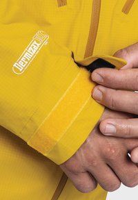 Gelbe wasserdichte Jacke aus strukturiertem Stoff, mit Klettverschluss am Ärmel und Logo-Detail, dazu eine Nahaufnahme einer Hand, die den Ärmel greift.