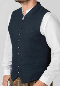Dunkelblaues Wollweste mit metallischen Knöpfen, Stehkragen mit blauer Stickerei und zwei vorderen Taschen. Über einem weißen Hemd getragen.