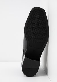Semelle de chaussure de ville noire avec motif de grip texturé, bout carré et talon bloc reposant verticalement contre un mur d’angle blanc.