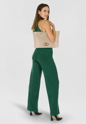 Sac cabas beige avec poignées chaîne dorée, orné d'un accent logo. Le modèle porte une tenue verte, présentant le sac de côté.