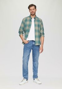 Groene en beige geprijsde button-up shirt over een witte t-shirt, gecombineerd met blauwe spijkerbroeken en witte sneakers. Eenvoudig ontwerp, casual katoen stof.