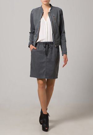 Giacca bomber in pelle grigia sopra una camicetta bianca, abbinata a una gonna grigia coordinata. L'outfit presenta una vita con coulisse e stivaletti alla caviglia.