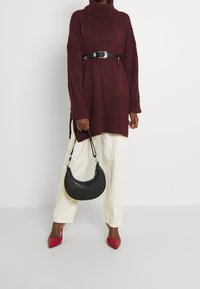 Maglione oversize color bordeaux con collo alto, orlo a coste, cintura nera con fibbia metallica, pantaloni beige a gamba larga e borsa nera. Scarpe con tacco rosso.