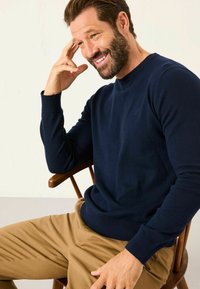 Navy knitted sweater with a round neckline and ribbed cuffs, paired with khaki pants. Model seated on a wooden chair, hand near his face.