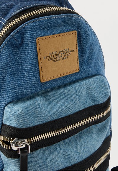 Τζιν σακίδιο πλάτης Marc Jacobs με ταμπελάκι από δέρμα καφέ που δείχνει το στυλ, την τοποθεσία και το έτος, με μαύρα διπλά φερμουάρ στην μπροστινή τσέπη.