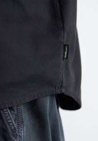 Zwarte shirt met een soepele stofstructuur en een licht gebogen zoom, voorzien van een klein Carhartt-logo op de zijkant.