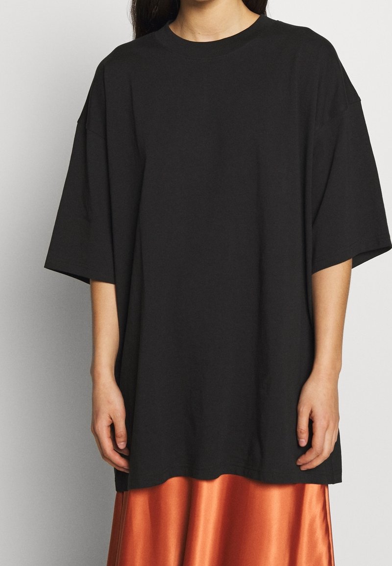 Schwarzes, übergroßes T-Shirt, kurze Ärmel, runder Halsausschnitt. Das Material scheint weich zu sein. Getragen über einem glänzenden, orangenen Satrock.