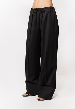 Kobieta w luźnych, czarnych spodniach o szerokich nogawkach z wiązaniem w talii oraz czarnych sandałach na otwarte palce, stojąca na białym tle.