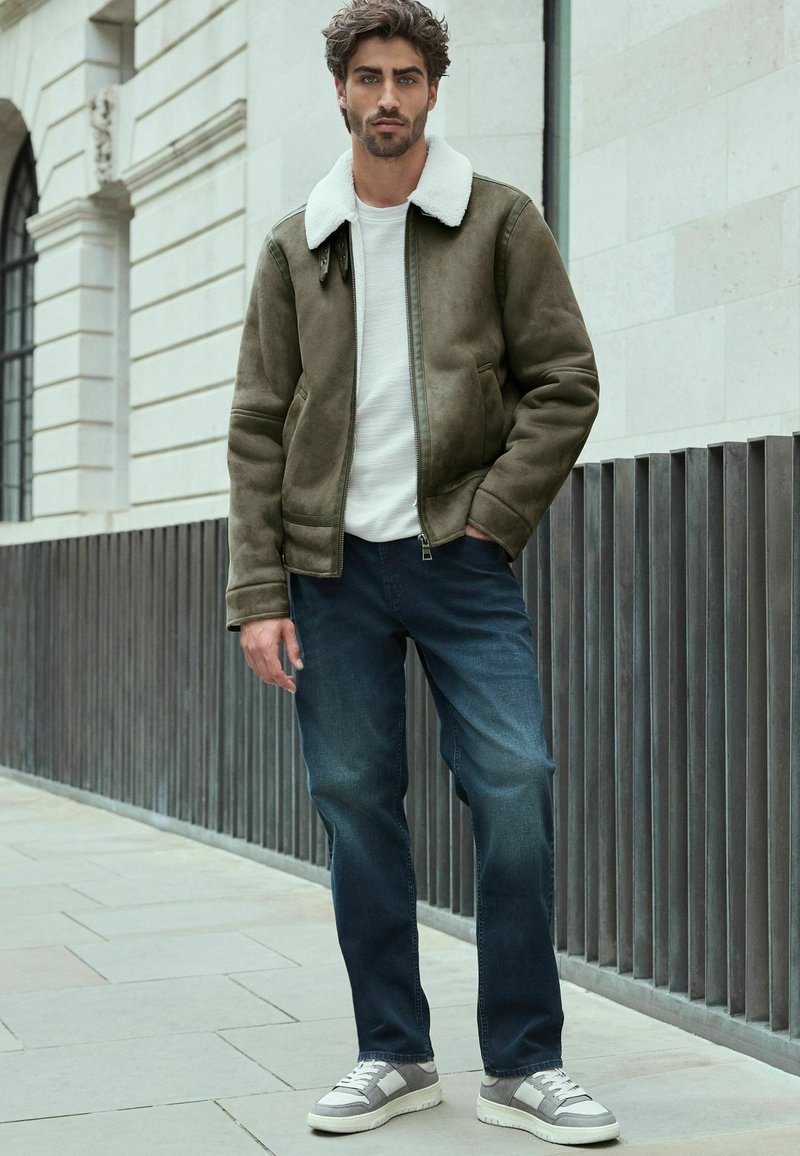 Chaqueta de ante verde oliva con un cuello de piel blanco, combinada con una camisa blanca y vaqueros azules. Zapatillas grises con rayas blancas.