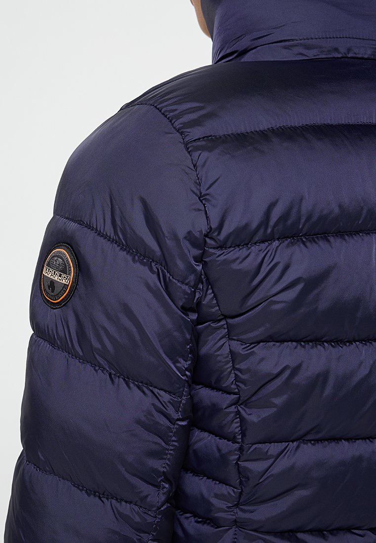 Marineblauwe pufferjack met een gequilt ontwerp, voorzien van een ronde logo patch op de linker schouder. Gemaakt van glad, lichtgewicht materiaal.