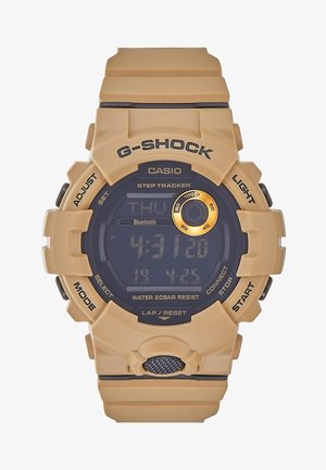 Montre digitale Casio G-Shock couleur beige avec podomètre, Bluetooth, étanche, boutons étiquetés et cadran rond avec accents noirs et dorés.