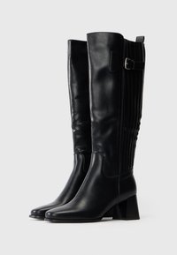 Par de botas de cuero negras hasta la rodilla con tacones cuadrados y detalle de hebilla lateral, mostradas sobre un fondo blanco liso.